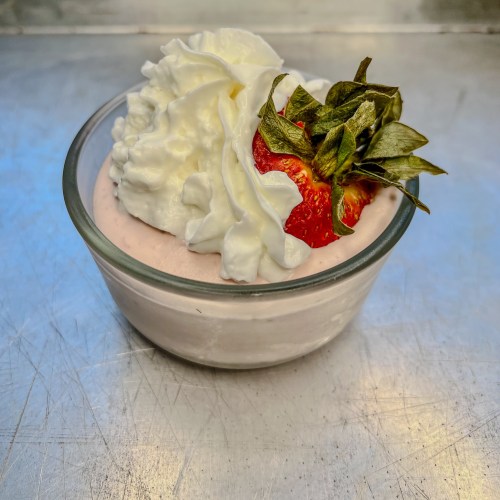 Strawberry mousse in a small dish with whipped cream and a strawberry on top, on a baking sheet