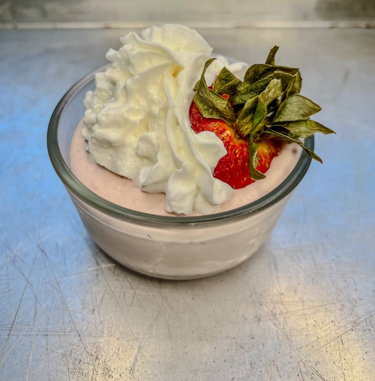 Strawberry mousse in a small dish with whipped cream and a strawberry on top, on a baking sheet