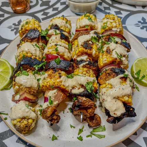 Street Corn Steak Kabobs on a plate with some lime, cilantro, and cotija crema drizzled on them and scattered all around