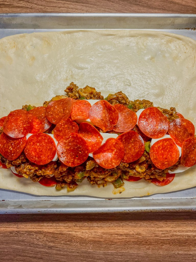 Pizza dough with mozzarella, pepperoni, and a meat mixture spread on it on a baking sheet