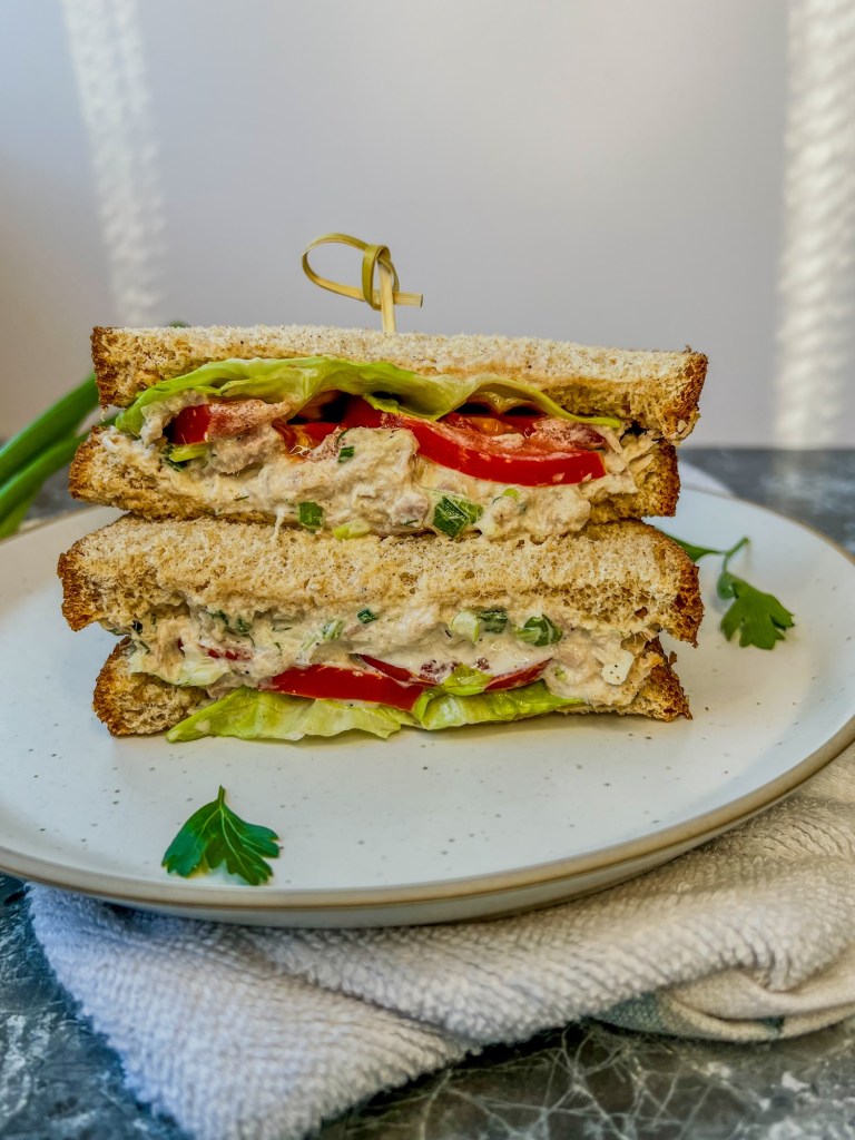 A tuna salad sandwich with tomato and lettuce that's been cut in half and stacked on a white plate