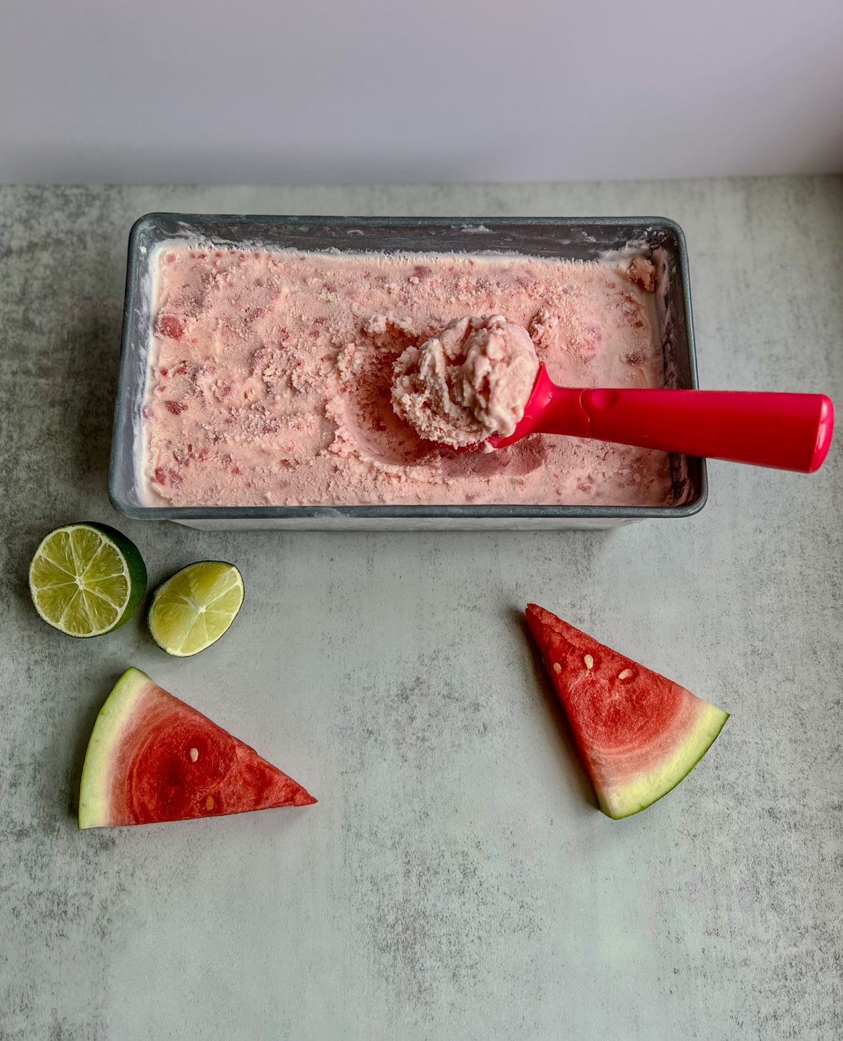 Watermelon Lime Ice Cream that's been scooped, sitting on a marble counter with lime and watermelon nearby