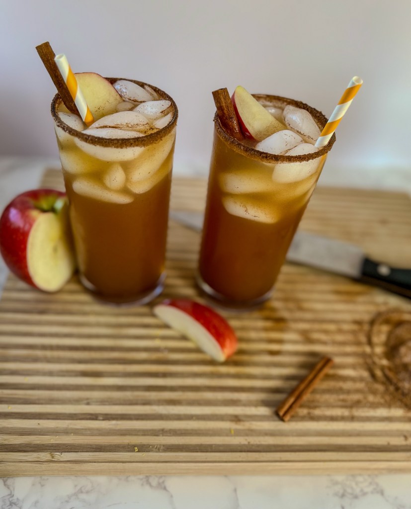 Two apple cider whiskey sour cocktails on a wooden cutting board with some apples, cinnamon sticks, and spices laying around
