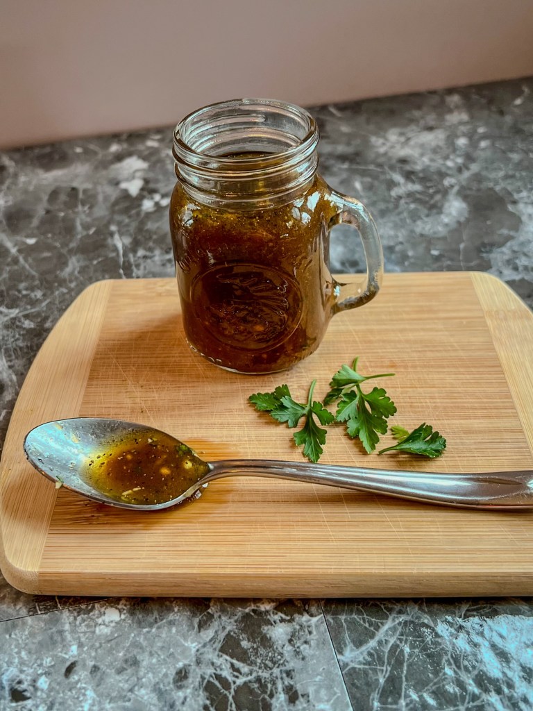 Honey balsamic vinaigrette in a small jar with a spoonful of it and some parsley next to it on a wooden cutting board