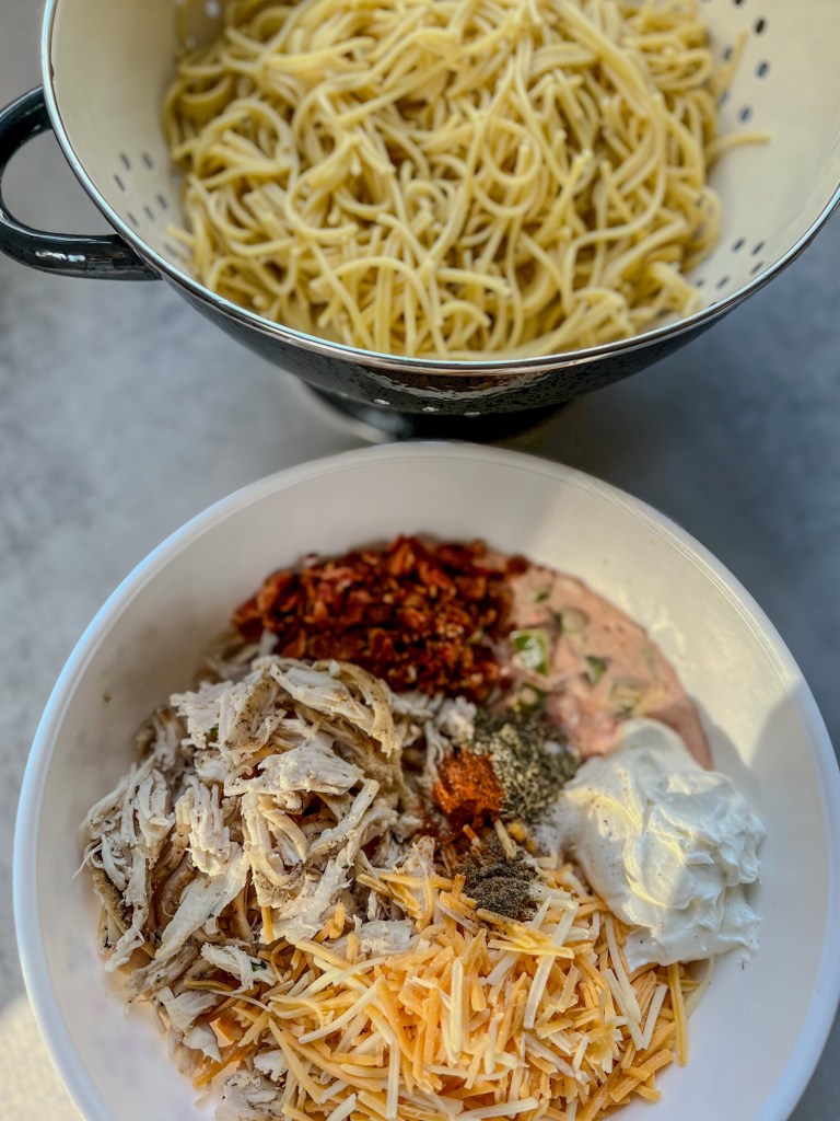 A large bowl filled with the filling for chicken spaghetti with a strainer filled with noodles behind it