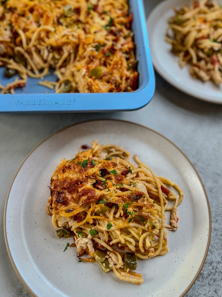 Baked chicken spaghetti fresh out of the oven on a plate with another plate and the baking dish with more behind it