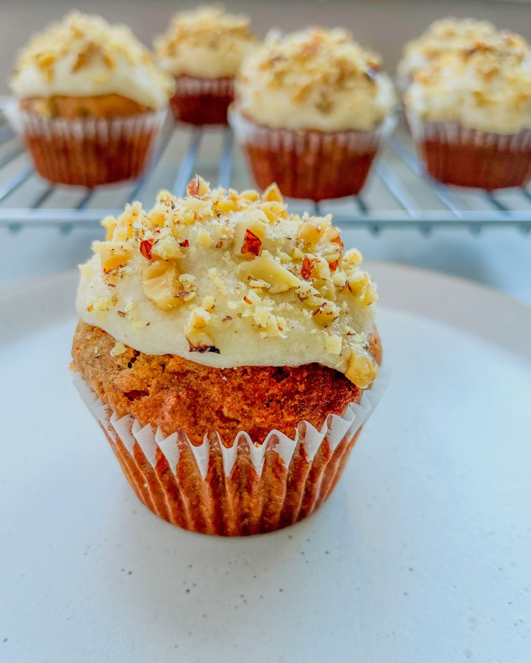 Banana walnut cupcakes with cream cheese frosting and toasted walnuts on top, sitting on a white plate with a rack of more cupcakes behind it