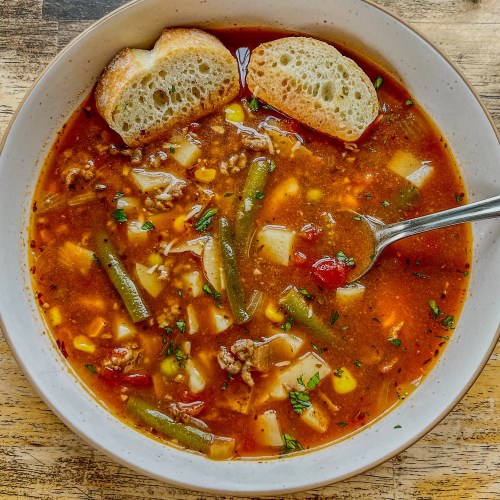 Beefy vegetable soup in a white bowl with some crispy bread and spoon inside the bowl, on a wooden table