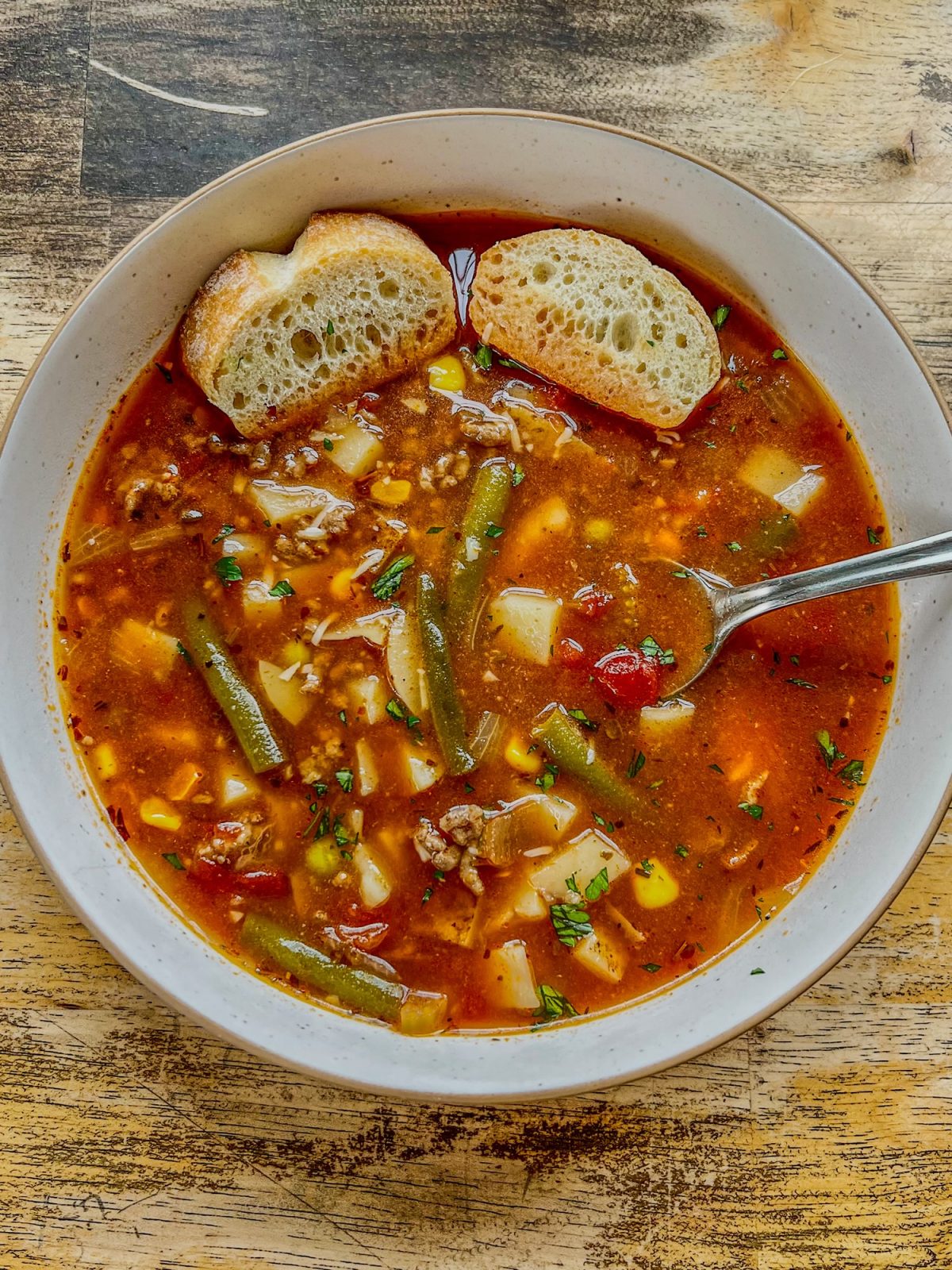 Beefy vegetable soup in a white bowl with some crispy bread and spoon inside the bowl, on a wooden table