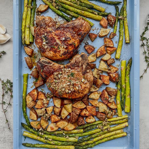 Bone-in pork chop sheet pan dinner on a baking sheet with some garlic butter scattered on top and fresh herbs and garlic nearby