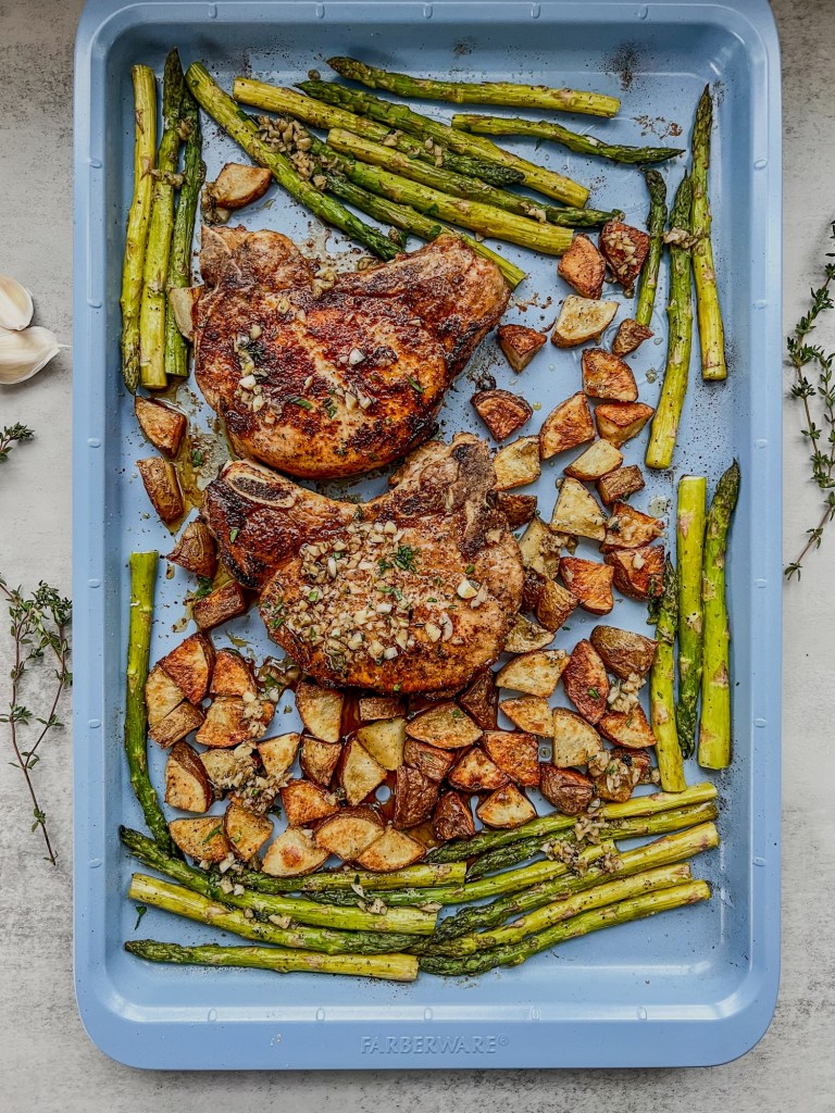 Bone-in pork chop sheet pan dinner on a baking sheet with some garlic butter scattered on top and fresh herbs and garlic nearby