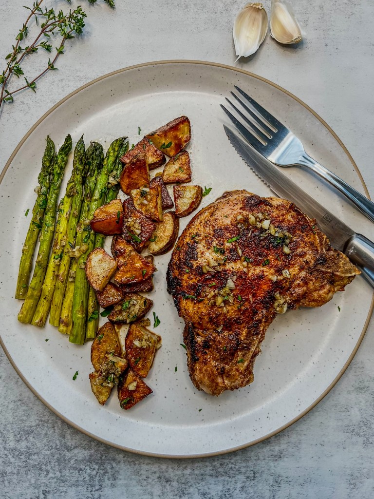 Bone-in pork chop sheet pan dinner on a white plate with some garlic butter drizzled over the top and a fork, knife, herbs, and garlic off to the side
