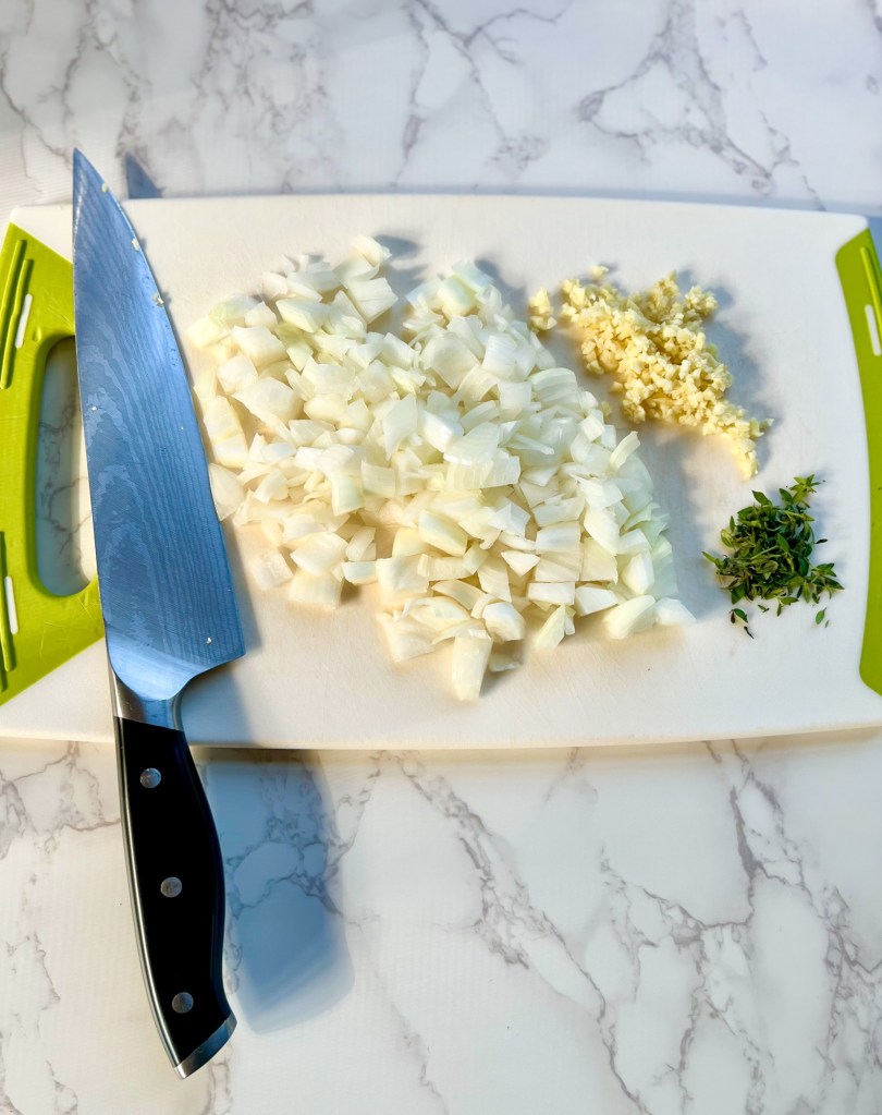 Onion, garlic, and thyme cut up on a cutting board with a knife nearby