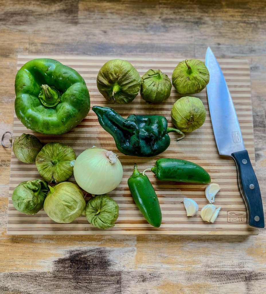 Peppers, garlic, onion, and tomatillos on a cutting board with a knife nearby