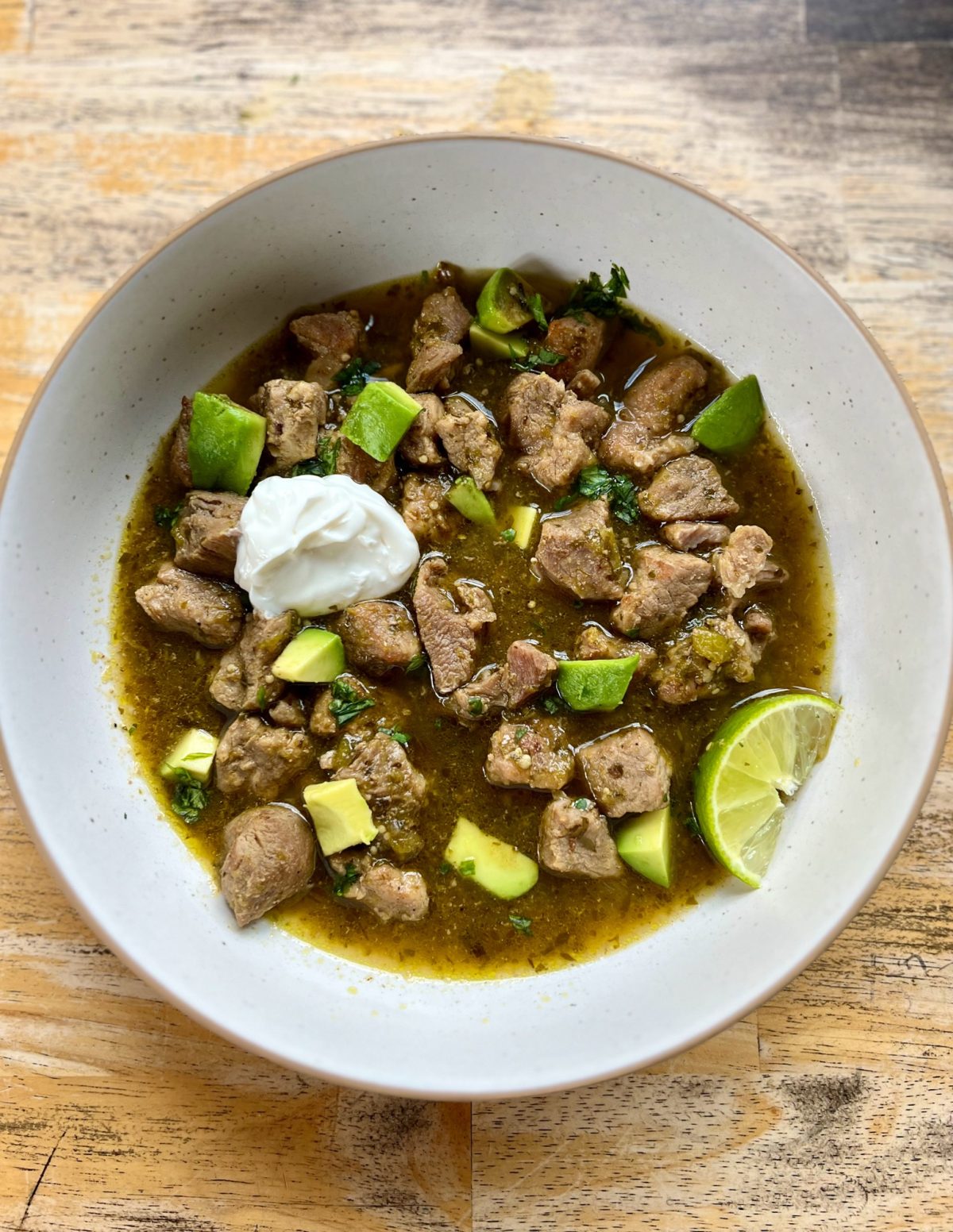 Chili Verde in a white bowl with sour cream, cilantro, avocado, and lime for garnish