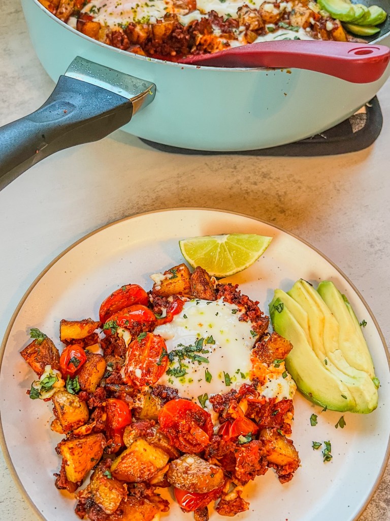 Chorizo hash on a white plate with some lime and avocado on the side, with a large skillet behind it