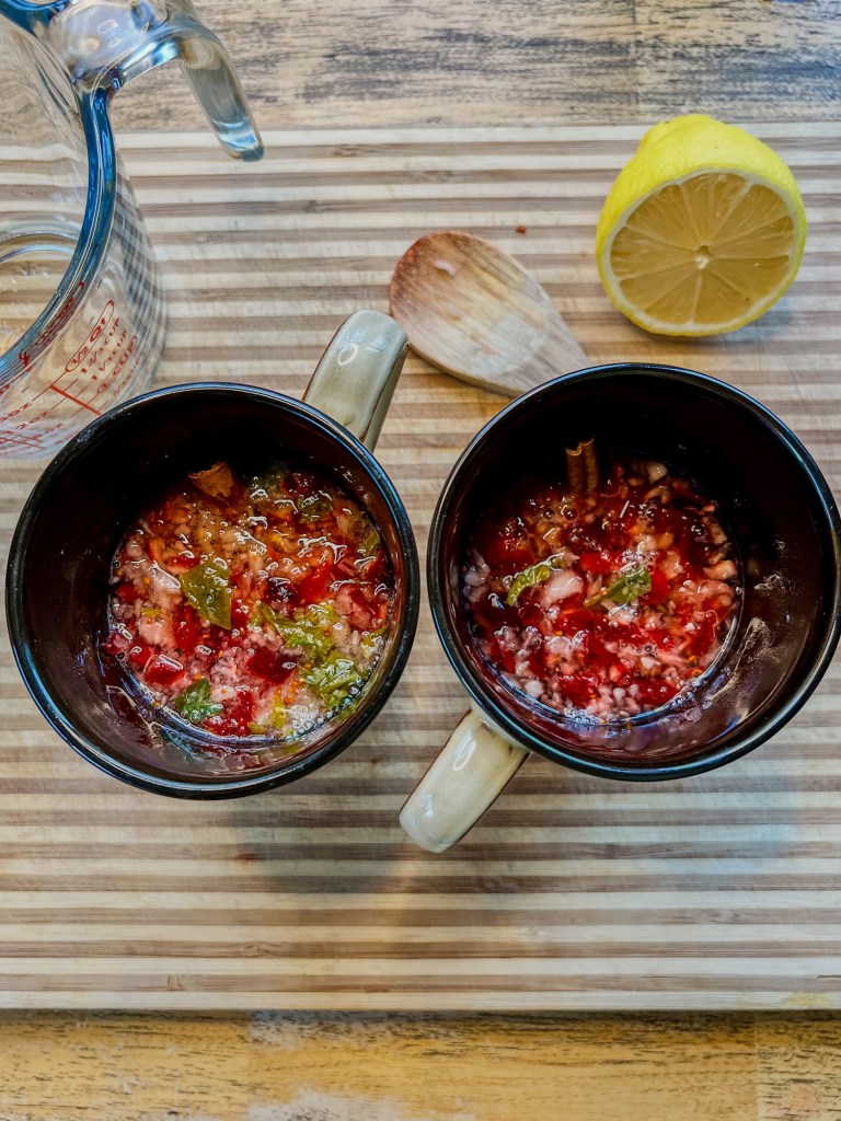 Cranberry, basil, hot water, and a cinnamon stick in some mugs with half a lemon behind