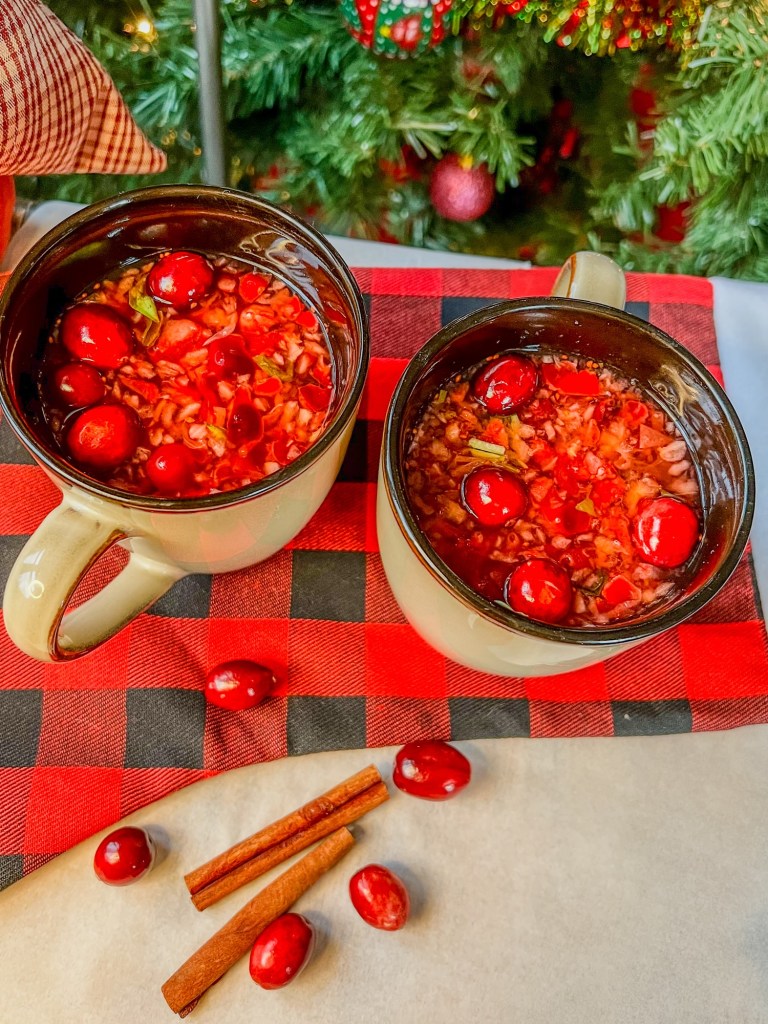 Two cranberry hot toddy drinks on a table with some cinnamon sticks and cranberries scattered around with a Christmas tree behind