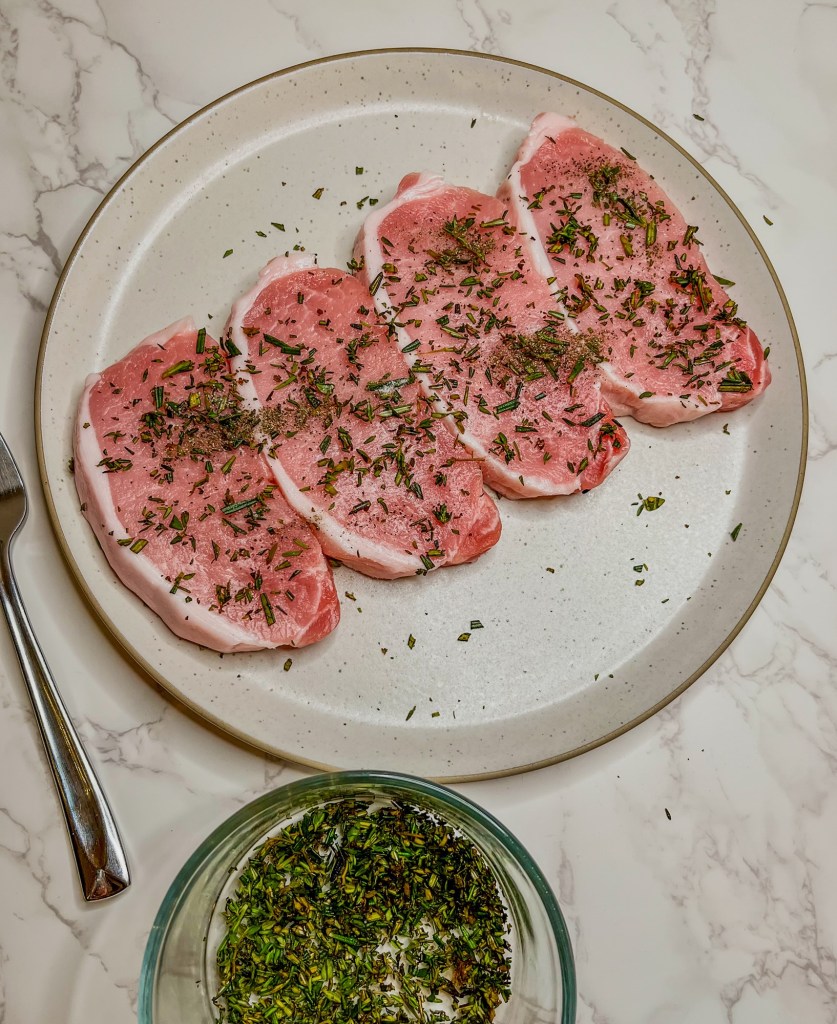 Pork chops on a white plate with some herbs sprinkled on them and a dish of herbs nearby