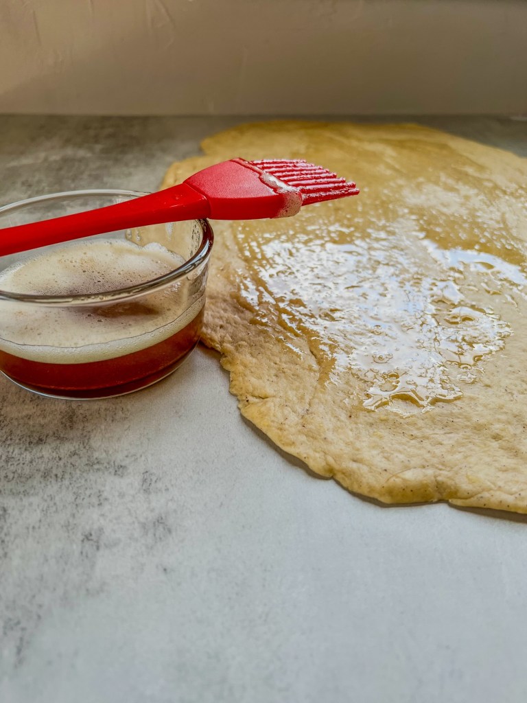 Brown butter in a dish with a silicone brush on top and cinnamon roll dough next to it