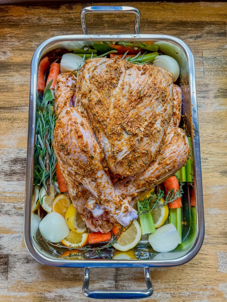 Turkey in a roasting pan that's been covered in herb spice butter with veggies and herbs scattered all around it, on a wooden table