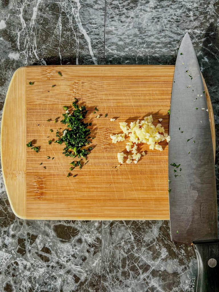 Garlic and parsley minced up on a wooden cutting board with a knife nearby