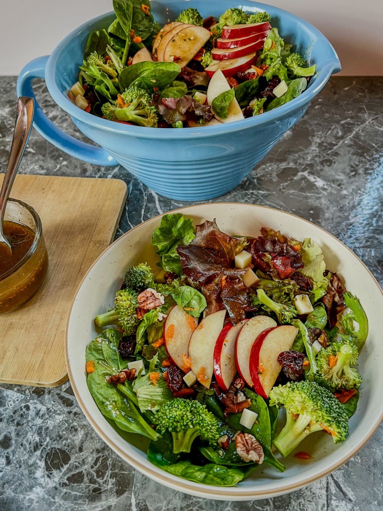 Apple salad in a bowl with a large bowl filled with more behind it with some honey balsamic vinaigrette off to the side