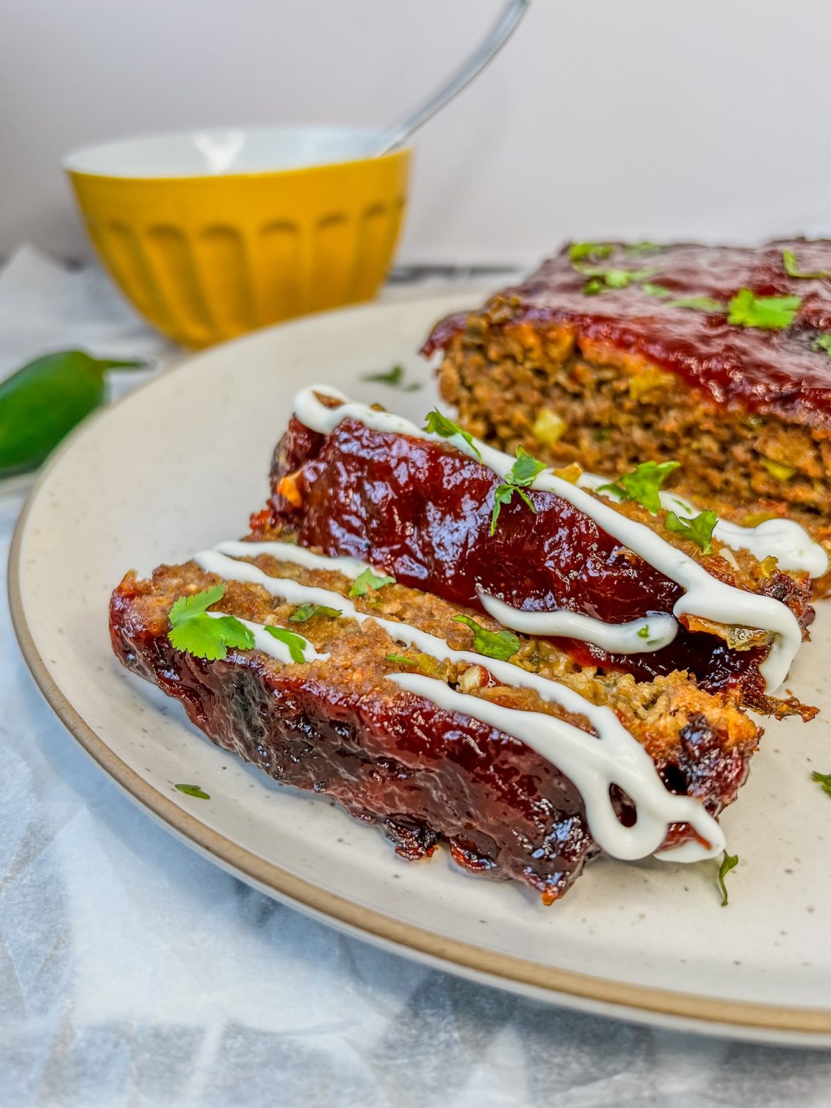 A jalapeño cheddar meatloaf fresh out of the oven, sliced, with a limey sour cream drizzled over the top, with some more sauce and a jalapeño off to the side