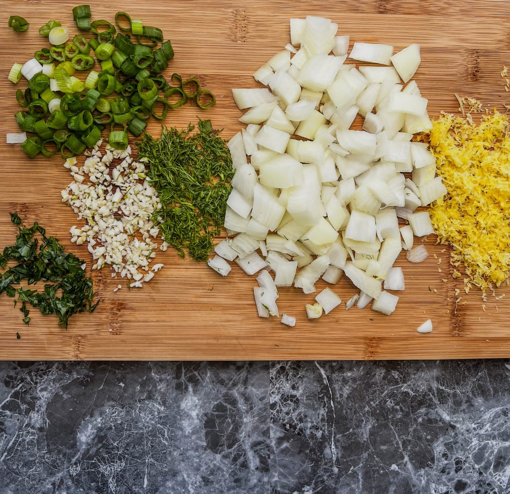 Onion, lemon zest, garlic, parsley, and dill chopped up on a cutting board