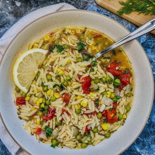 Lemon orzo soup in a bowl with some more soup, fresh herbs, and lemon nearby