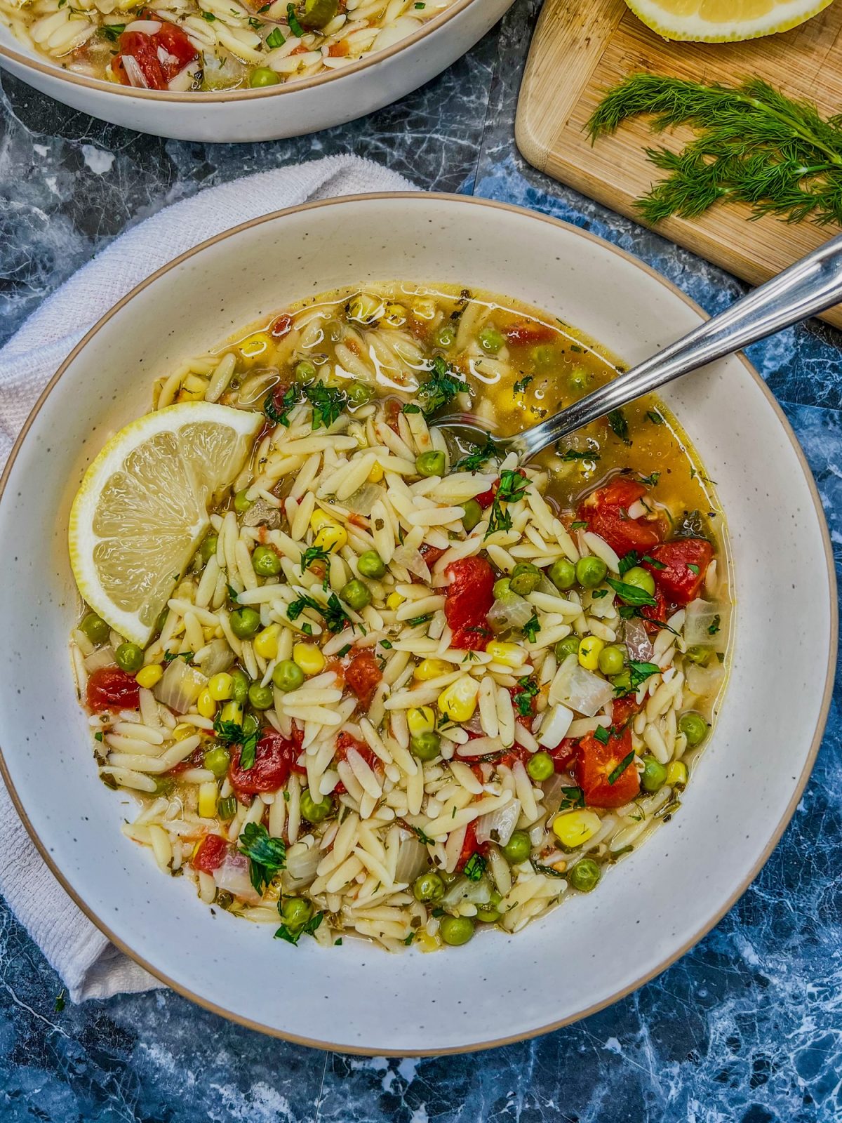 Lemon orzo soup in a bowl with some more soup, fresh herbs, and lemon nearby