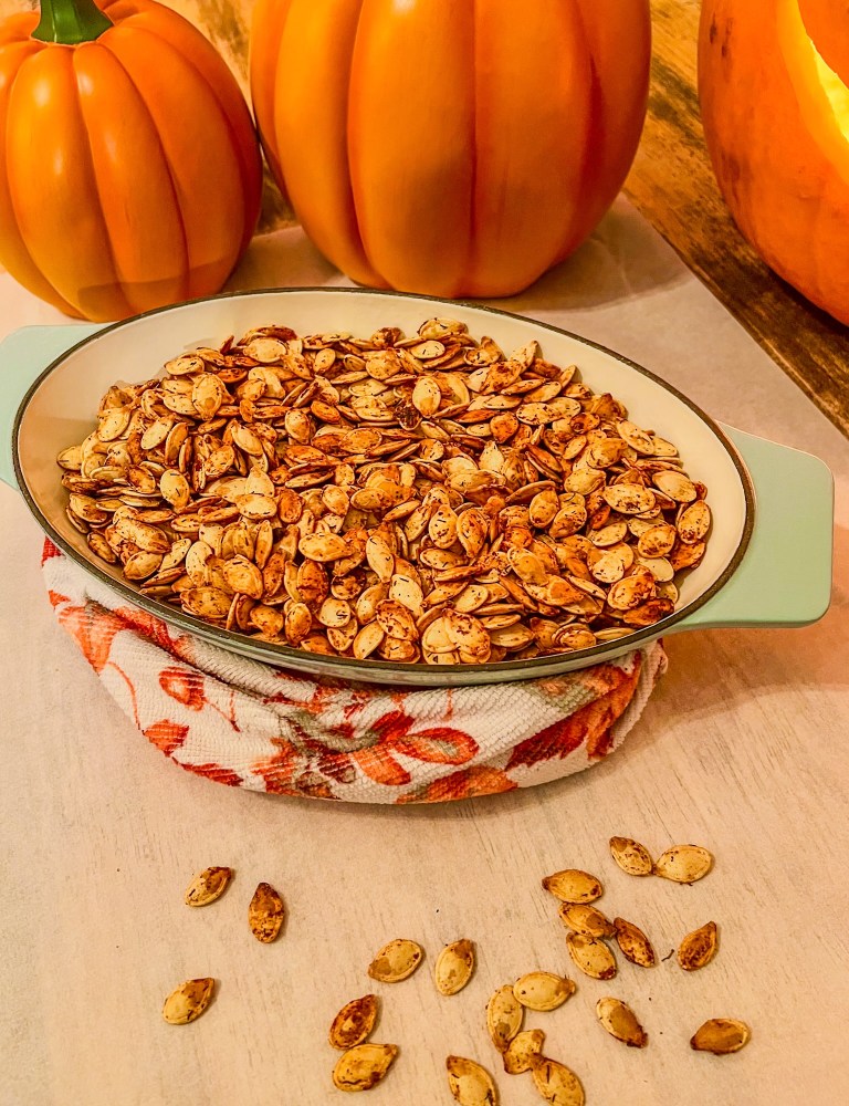 Lemon Pepper Pumpkin Seeds in a glass dish with some pumpkins behind and some seeds scattered around