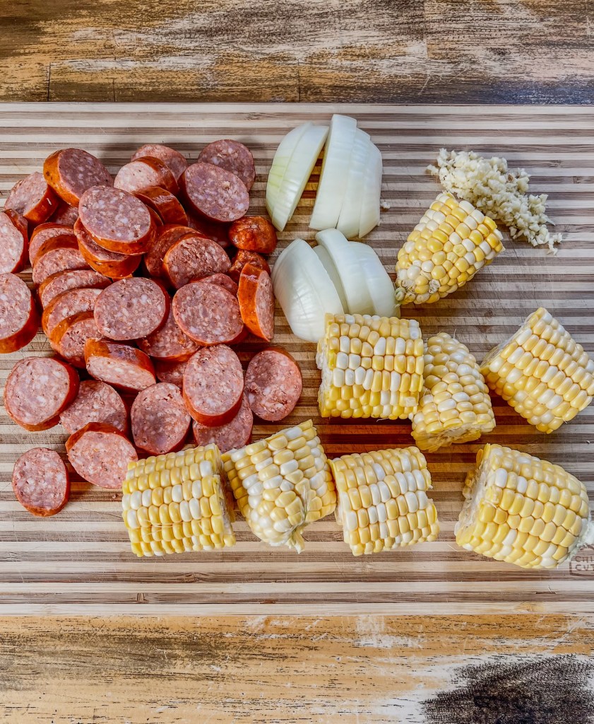 A cutting board with corn, onion, sausage, and garlic cut up on it