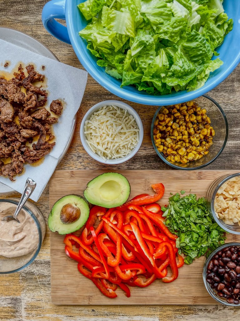 Avocado, steak, lettuce, black beans, bell pepper, cilantro, wonton strips, pepper jack cheese, roasted corn, and chipotle lime dressing spread out on a table for southwest steak salad