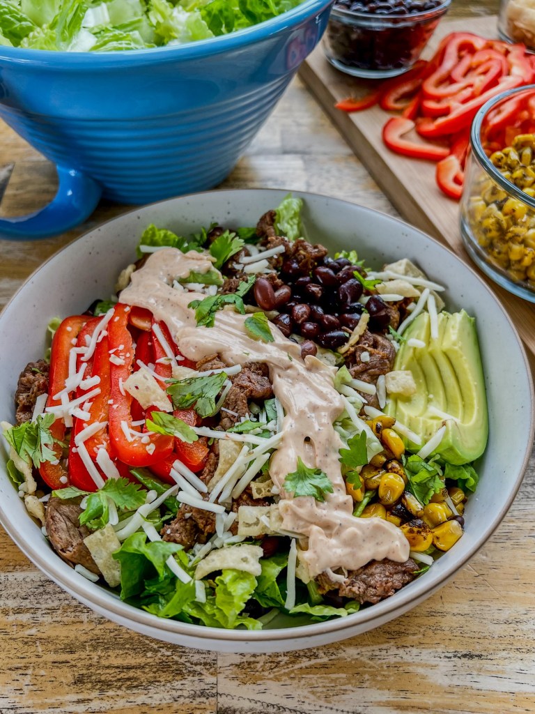 Southwest steak salad in a bowl with a creamy chipotle lime dressing over the top and all the fixings scattered around