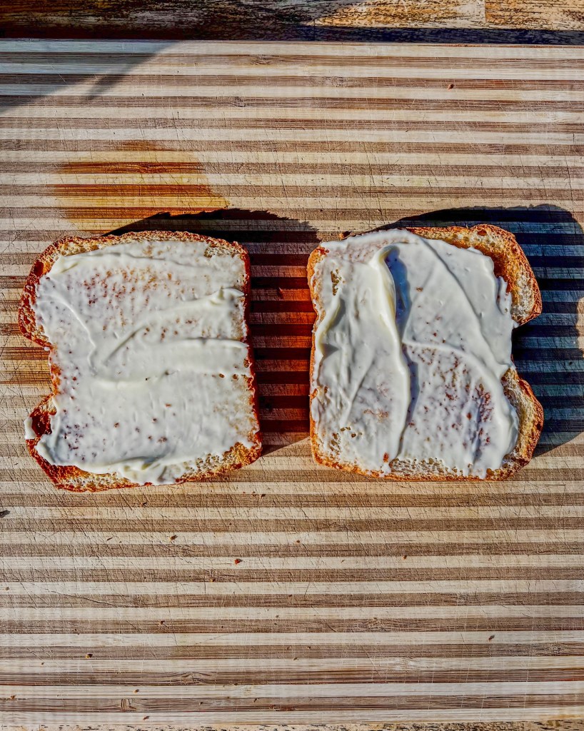 Two pieces of toast with mayo on them on a wooden cutting board