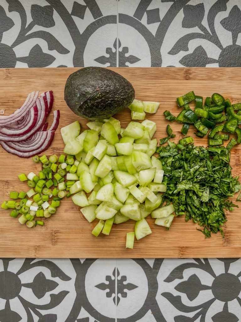 Red onion, cucumber, cilantro, green onion, jalapeño, and avocado on a wooden cutting board