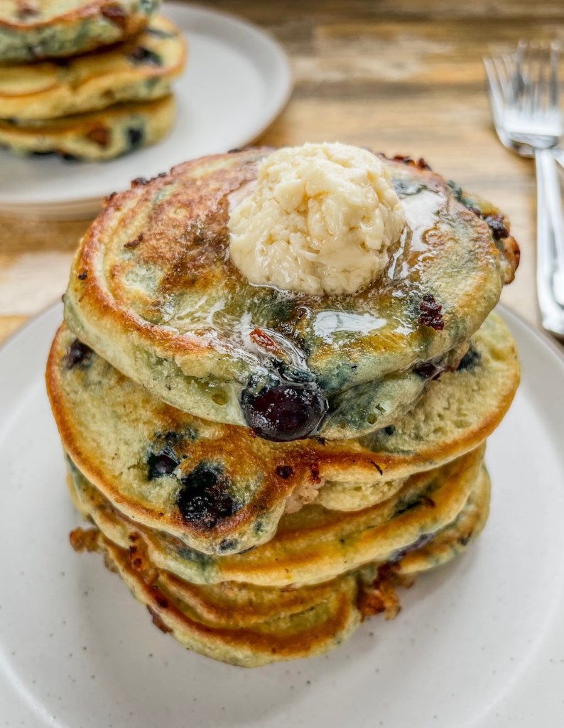 Blueberry sausage stuffed pancakes in a stack with maple butter on top with another stack behind and some silverware off to the side