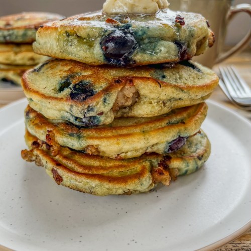 Blueberry sausage stuffed pancakes with maple butter on top and maple syrup, with some more pancakes and coffee behind