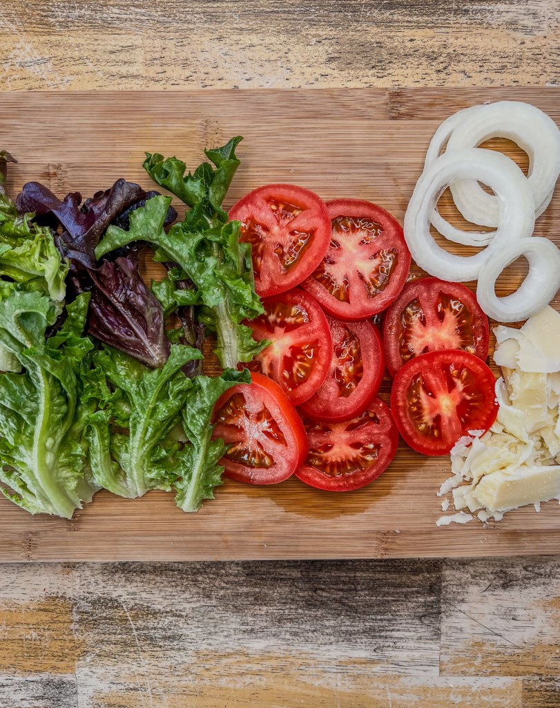 Lettuce, tomato, onion, and parmesan cheese prepped on a cutting board