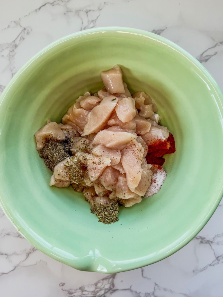 Chicken breast cut into chunks in a mixing bowl with spices and oil