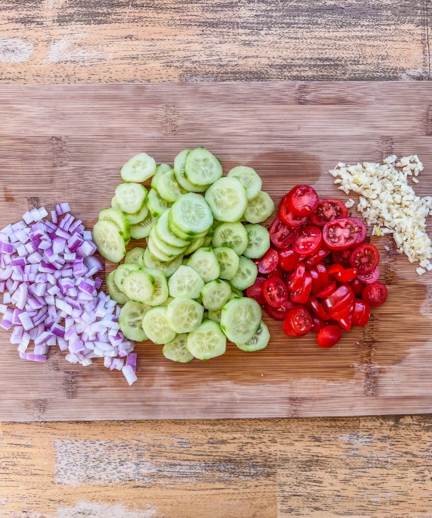 Onion, garlic, tomato, and cucumber cut up on a wooden cutting board