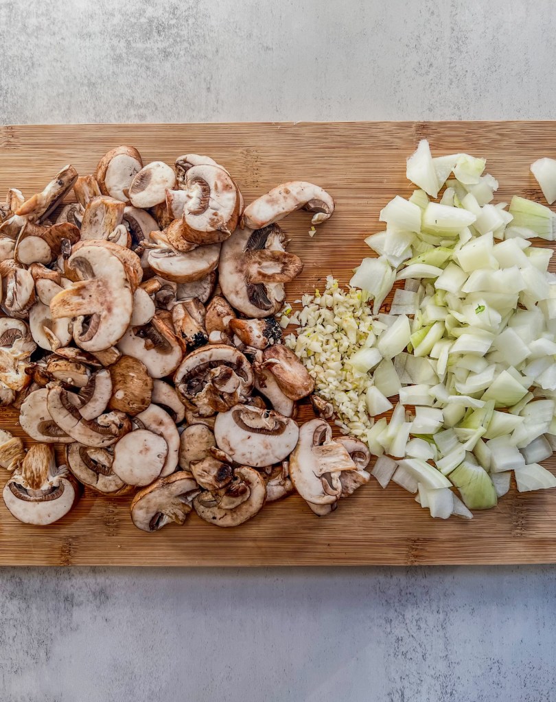 Baby bella mushrooms, onions, and garlic cut up on a cutting board