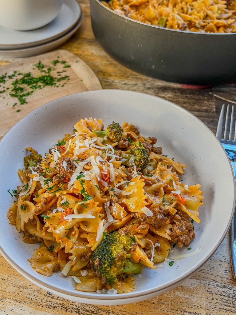 Creamy tomato beef pasta in a bowl with some fresh parsley and parmesan on top with more pasta in a skillet behind