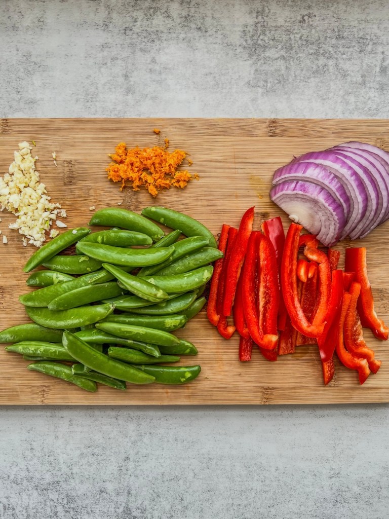 Red pepper and onion, sugar snap peas, garlic, and orange zest prepped on a cutting board