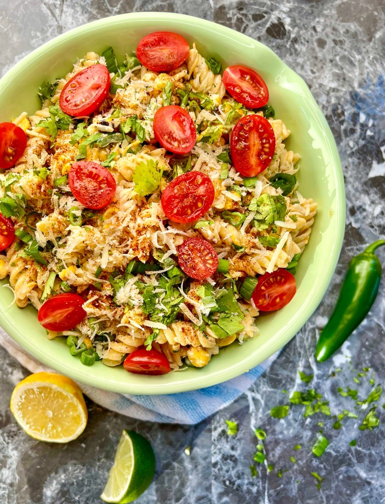 Street corn pasta salad in a bowl with some lemon, lime, jalapeño, and herbs scattered around