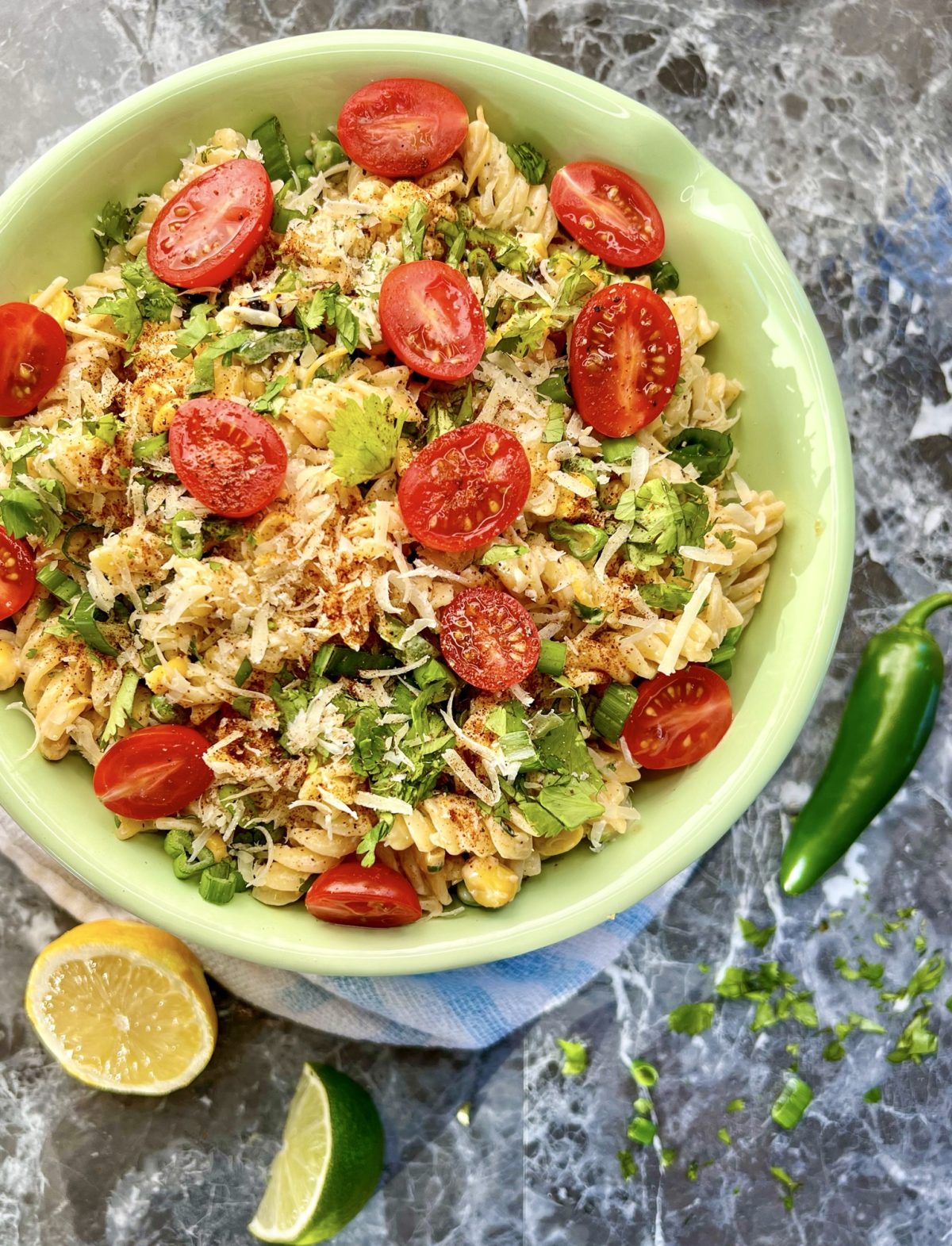 Street corn pasta salad in a bowl with some lemon, lime, jalapeño, and herbs scattered around