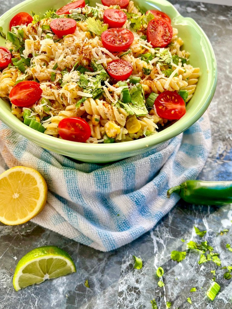 Street corn pasta salad in a large bowl with some jalapeño, lemon, lime, and herbs scattered around