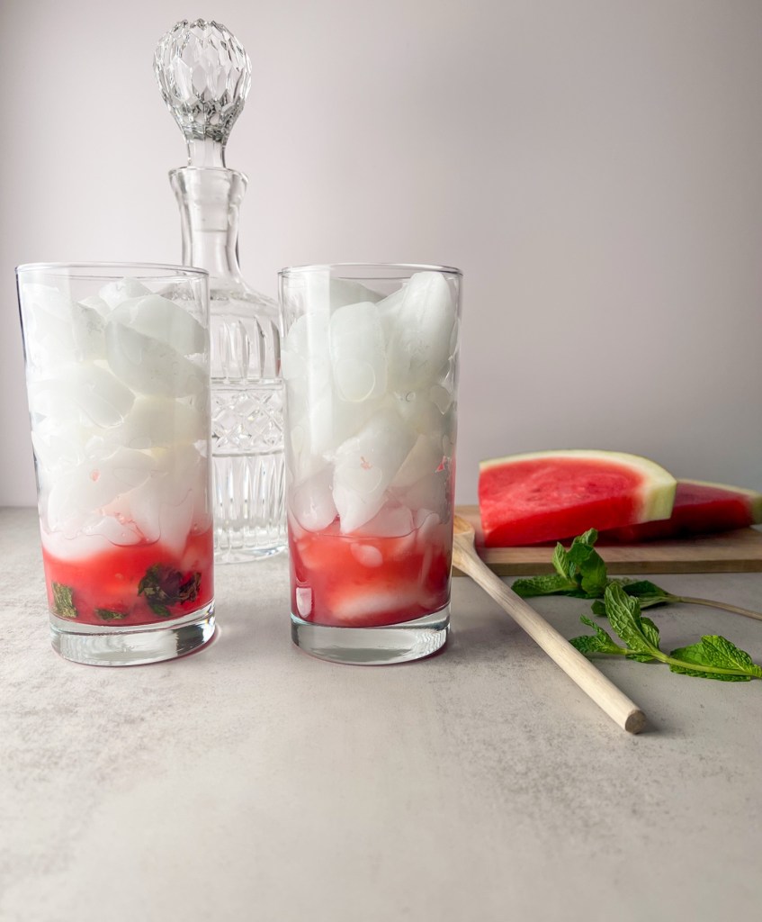 Two glasses filled with ice and muddled mint leaves and watermelon cubes.