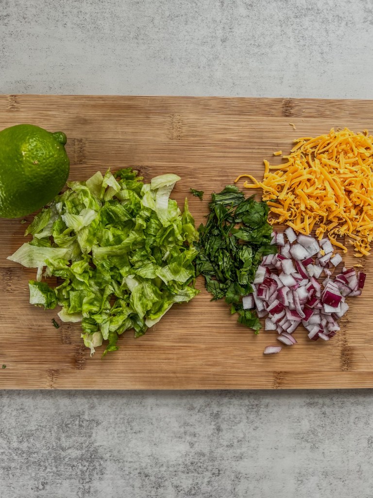 Lettuce, red onion, cilantro, shredded cheddar, and a lime on a wooden cutting board